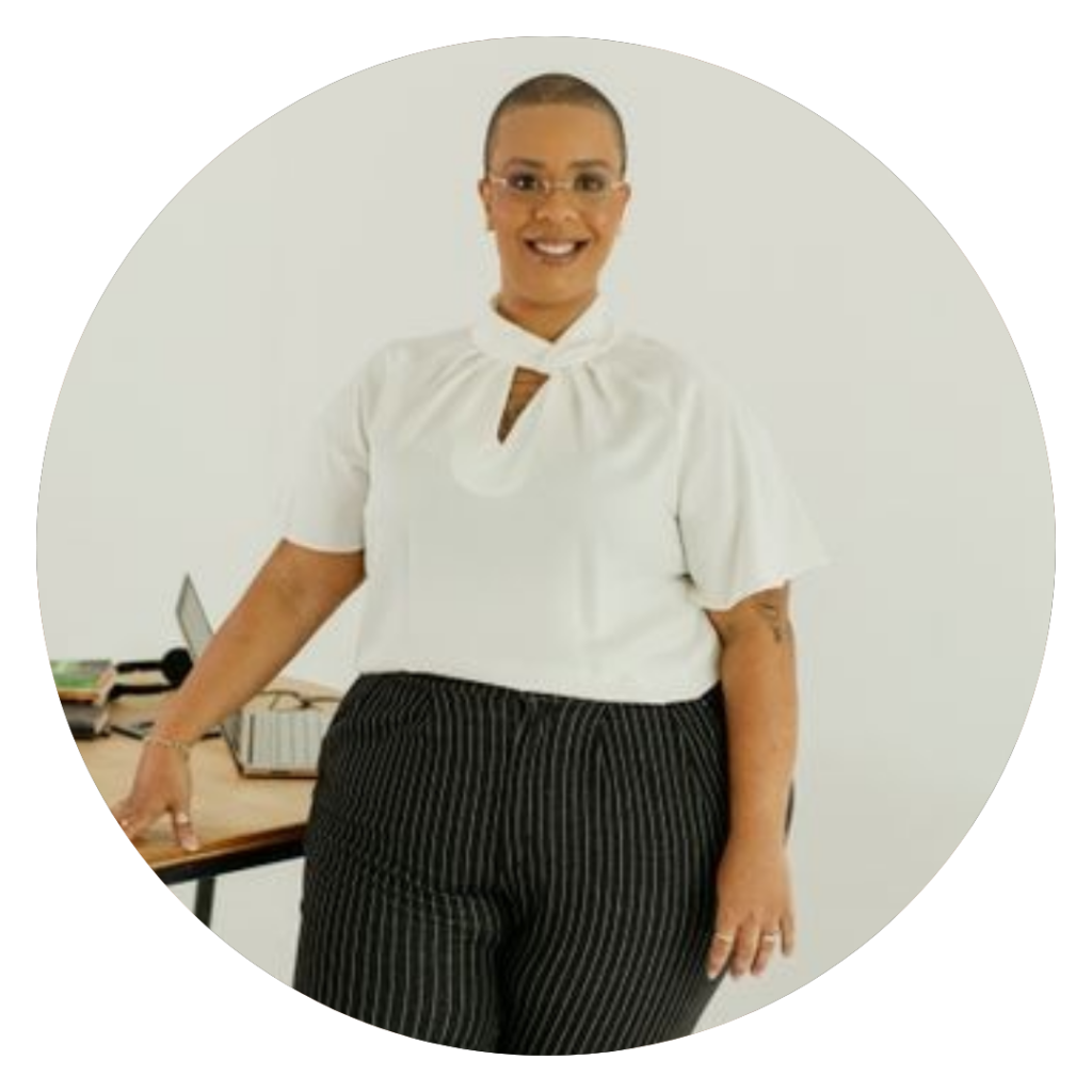 Foto da thais de pe com mesa ao fundo, utilizando uma blusa branca e uma calça preta